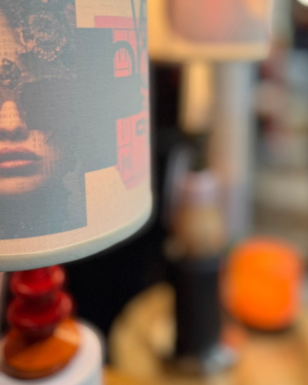 A close-up of a lampshade featuring a graphic print of a woman's face with gears and pink architectural shapes, with a blurred background showing other home decor items.