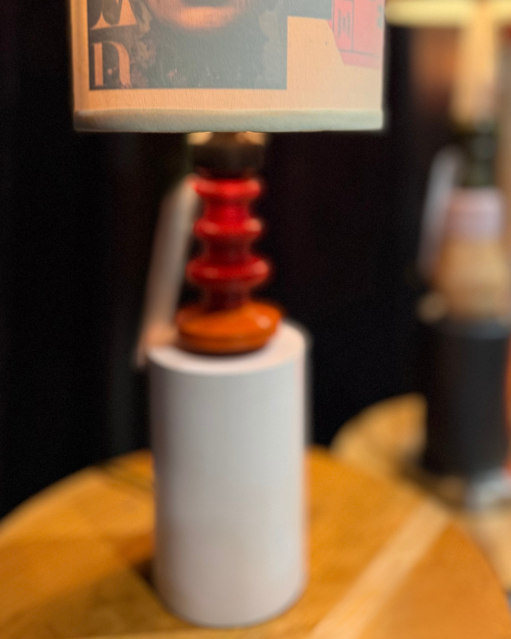 A close-up of a table lamp with a white cylindrical base, a red tiered stem, and a beige lampshade featuring a graphic print, set on a wooden surface.