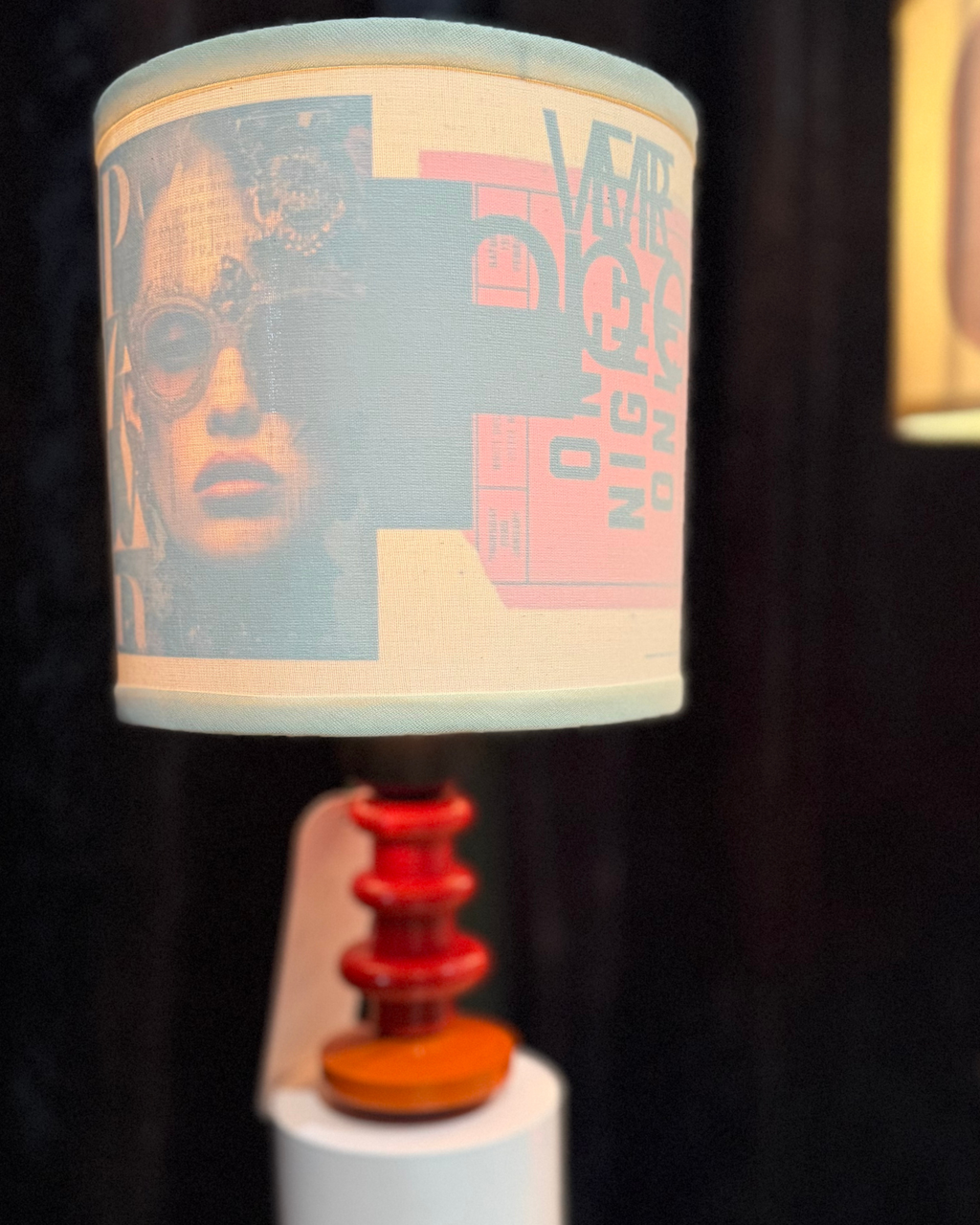 A table lamp with a cylindrical drum shade featuring a graphic collage print of a woman's face and the text 'Vampire Night Out', supported by a tiered red and orange wooden base on a white pedestal.