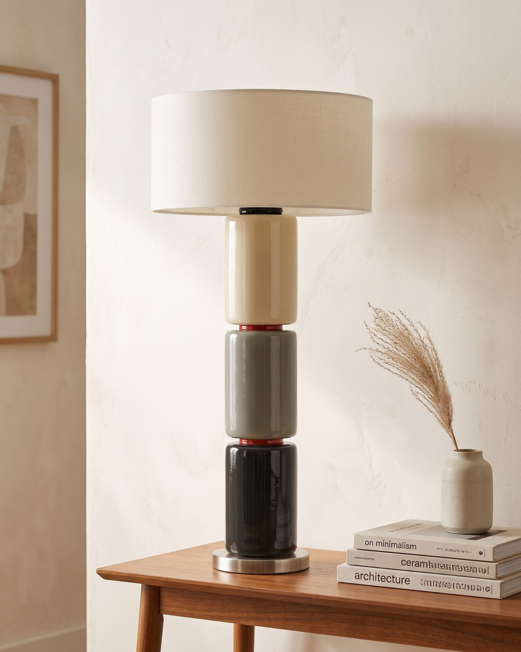 A modern stacked table lamp with a cream drum shade and cylindrical base sections in cream, gray, black, and dark red, displayed on a wooden side table in a warm interior setting.