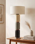 A modern stacked table lamp with a cream drum shade and cylindrical base sections in cream, gray, black, and dark red, displayed on a wooden side table in a warm interior setting.