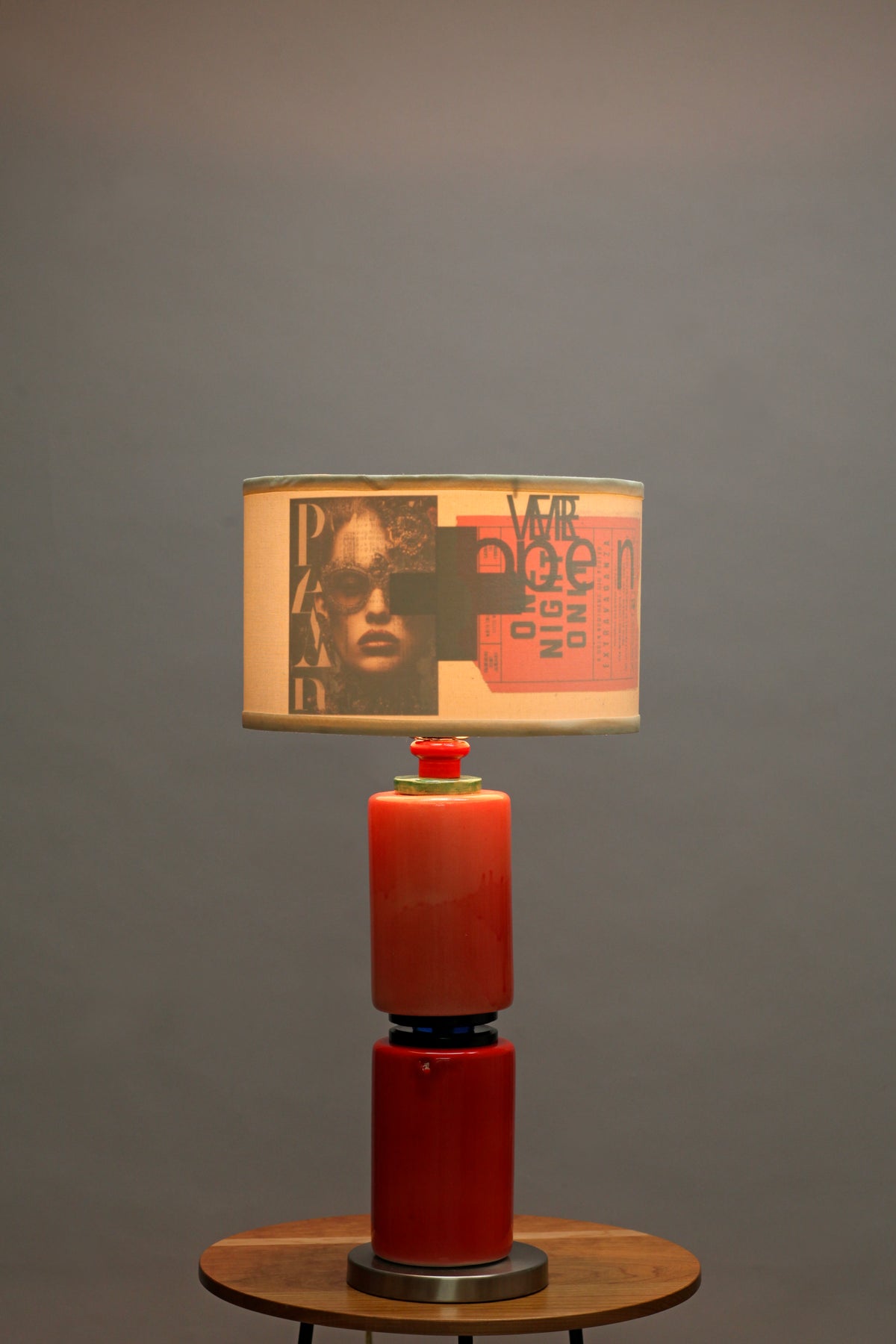 A modern table lamp with a cylindrical red glass base and a drum shade printed with a vintage-style poster design, shown lit against a neutral gray background.