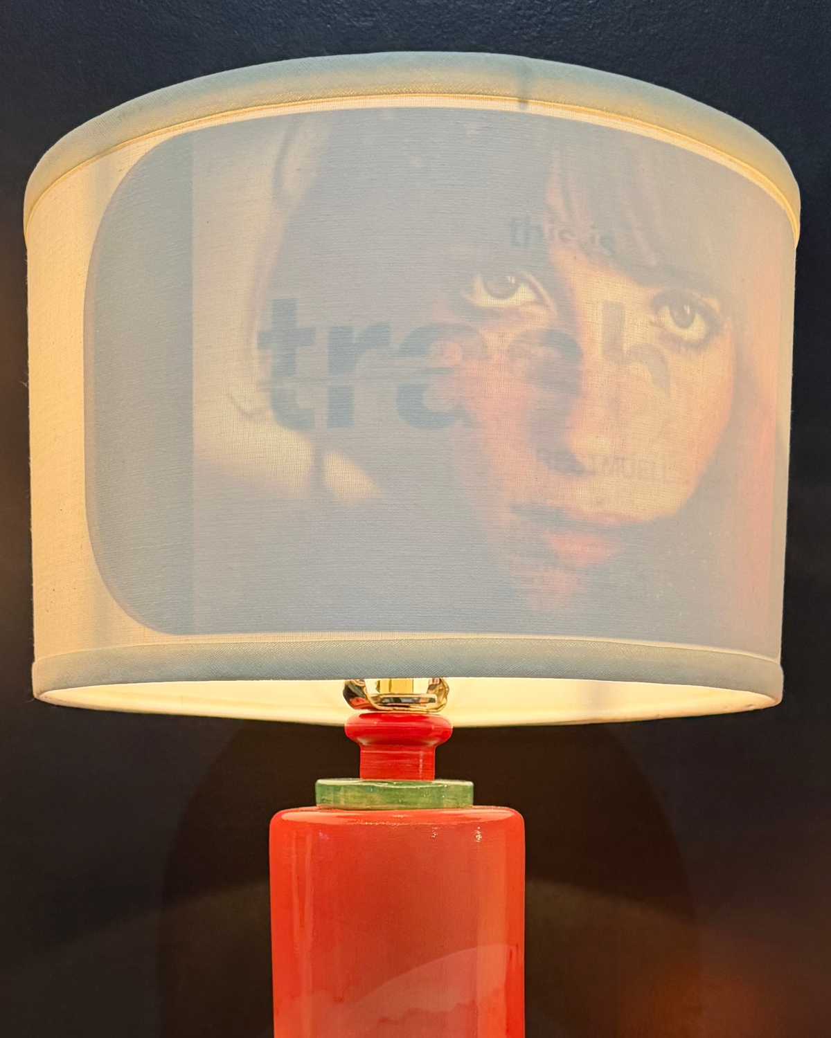 A table lamp with a cylindrical red-orange base, a green collar detail, and a drum shade made from translucent printed fabric featuring a face and the word 'trash'.