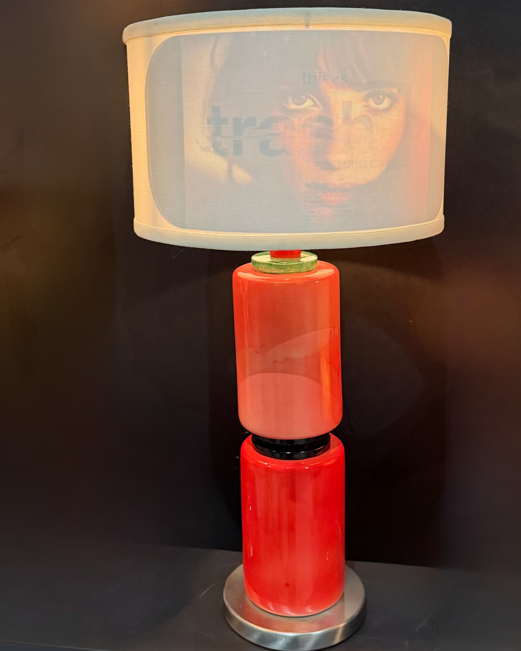A modern table lamp with a glossy red cylindrical base and a white drum shade featuring a printed portrait of a woman, set against a dark background.