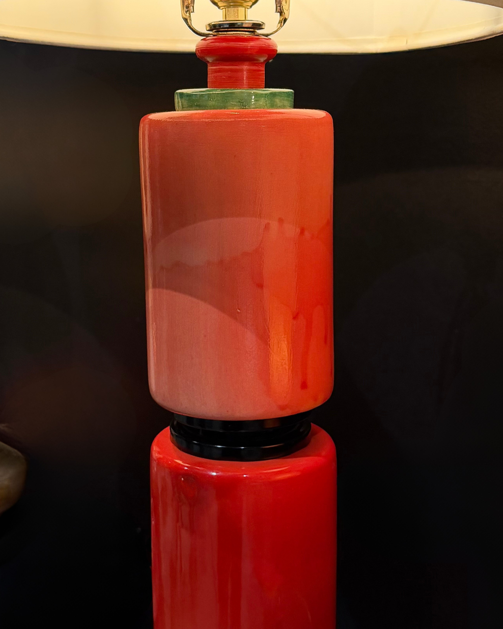 A close-up of a table lamp with a glossy, stacked ceramic base in vibrant red and orange tones, featuring a cream-colored lampshade at the top.