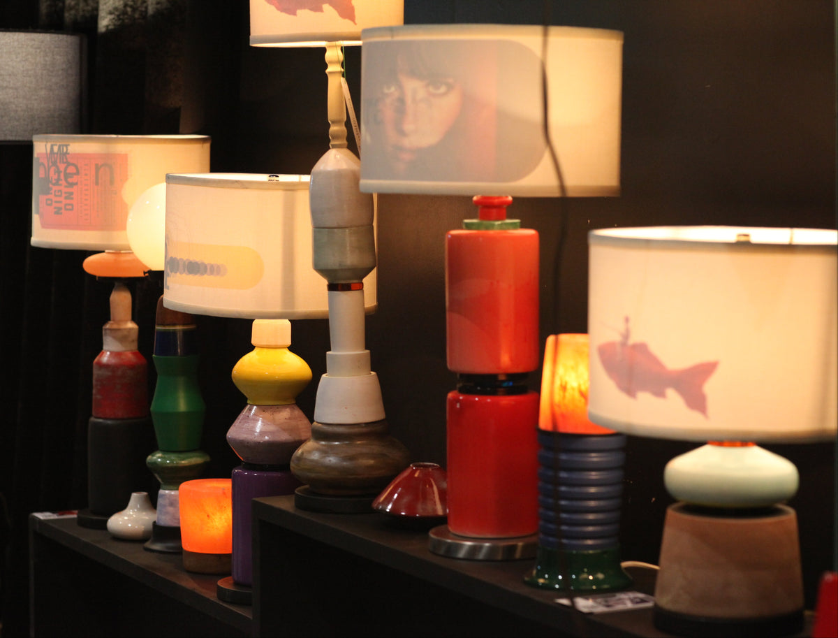 A collection of unique, colorful sculptural table lamps displayed on a dark shelf in a dimly lit room. The lamps feature various modern designs, including stacked cylindrical bases in red, white, and black, and white drum shades with printed graphics.