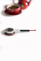 A modern, minimalist tea strainer with a white body, black handle, red tip, and dark wood base, resting on a white surface with a blurred red teacup in the background.