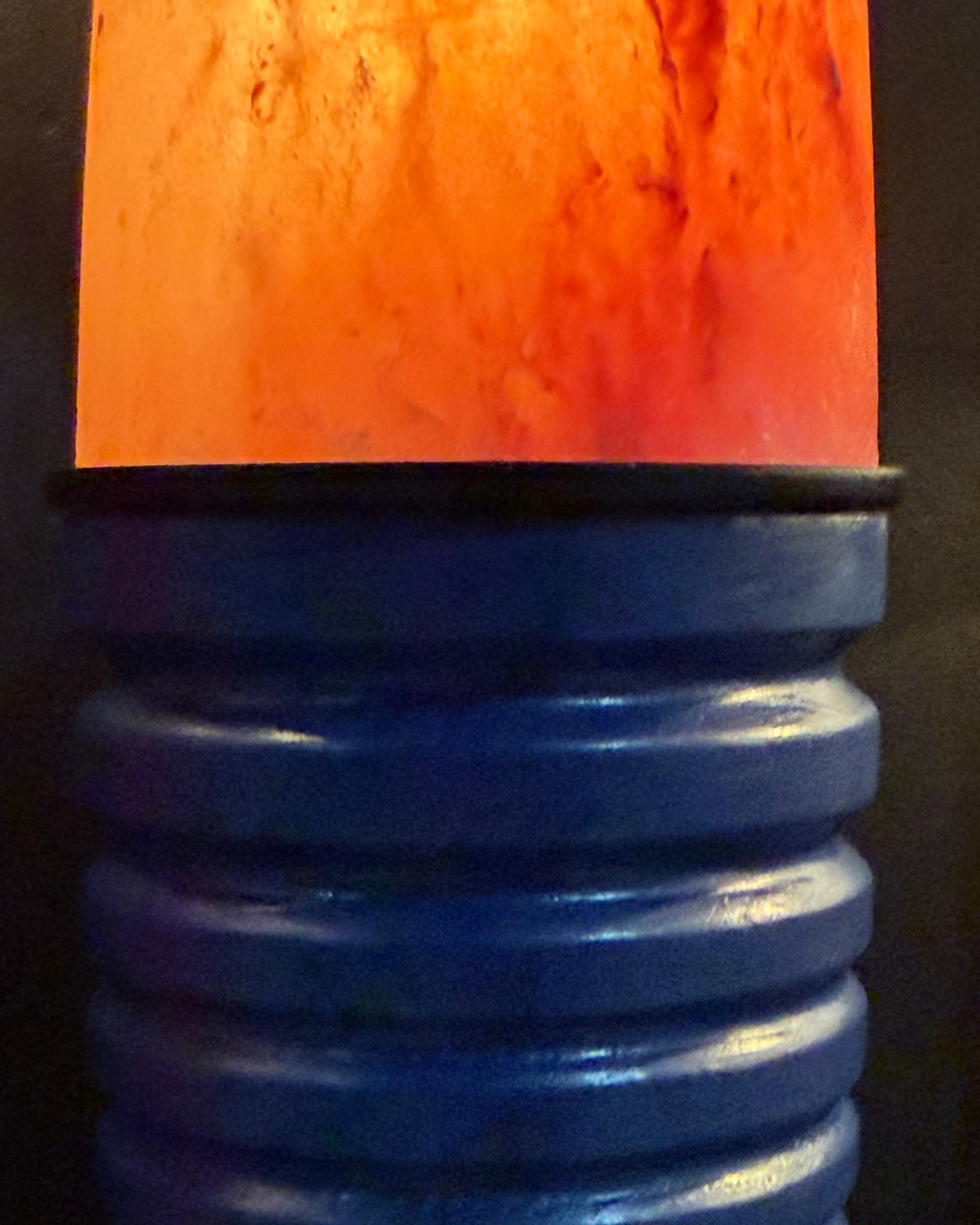 A close-up of a decorative lamp or vase-like object with a glowing orange-red translucent top section and a ribbed blue base against a dark background.
