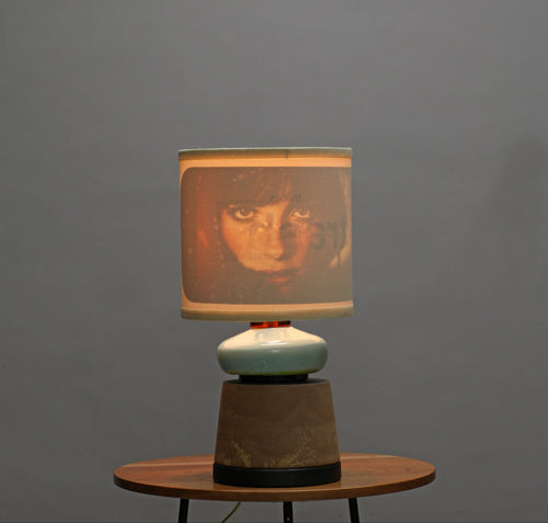 A table lamp with a square fabric shade featuring a printed portrait, resting on a small round wooden side table against a plain gray wall.
