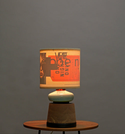 A table lamp with a cylindrical drum shade featuring a collage print of black, orange, and cream tones, resting on a dark wood base and a small round wooden side table against a solid gray wall.